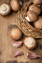 Aerial view of portobello mushrooms in wooden bowl on wooden table with more mushrooms and garlic, vertical