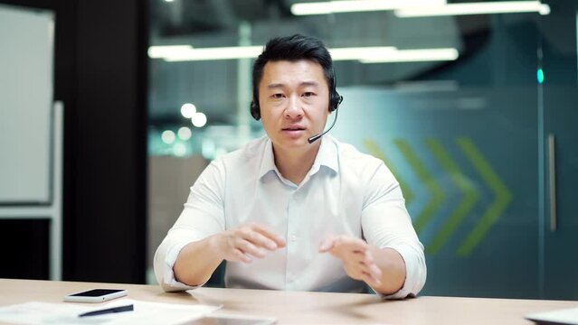 Asian business man manager holds online meeting conference remotely by video call using a webcam, headset. Office worker office with laptop computer. Talk look at camera view