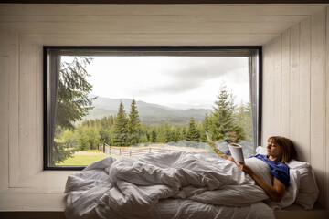 Woman reads a book while lying covered with blanket on a window sill with great view on mountains, resting in house on nature