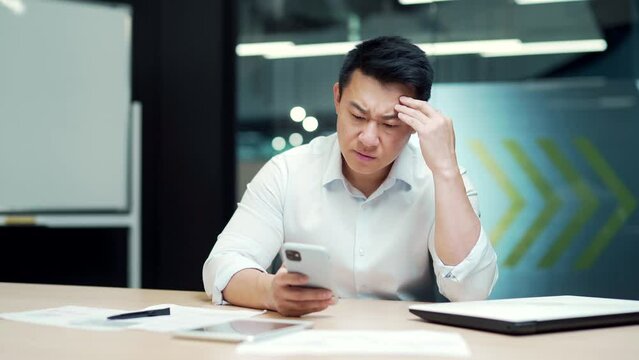 worried asian office worker employee reading bad news using smartphone while sitting at workplace. Upset business man entrepreneur investor looking at mobile phone sad looking at financial results