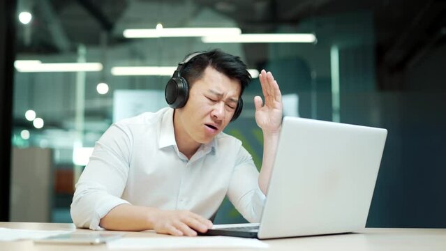 excited asian businessman cheering for game looking at laptop computer screen in office Male employee in headphones fail in sports betting fan watches an online match failure bad result soccer