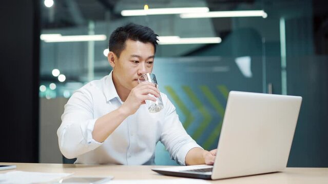 Asian employee man works in the modern office at the workplace sitting at the laptop computer drinking clean water from a glass. Business man, worker, quenches thirst Indoors