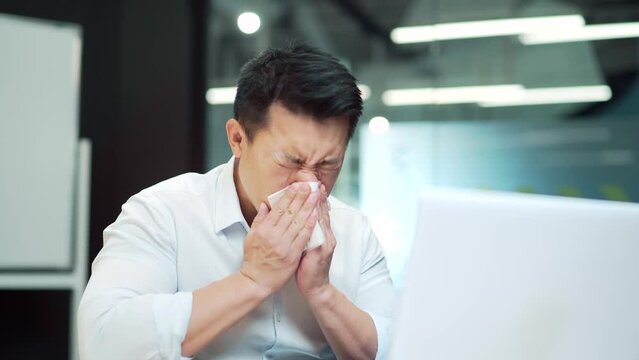 Handsome asian male worker suffers from allergies, sneezes with a runny nose. An employee coughs a cold or flu sitting at work in the office. Indoor. Feeling unwell in a business man suffering a cold