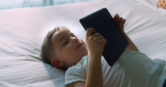 Cute smiling child, kid boy using digital tablet technology device lying on bedroom alone. Small child hold pad computer surfing internet play game at home