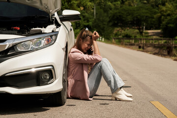 Depressed view of a young woman sitting on the road near a broken car and talking to a mechanic on the phone.