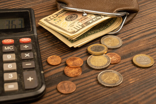 Dollar banknotes, euro coins, a vintage brown leather wallet and a calculator on a wooden surface.