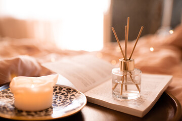 Bamboo sticks in bottle with scented candles and open book on wooden tray in bed closeup. Home cozy aroma.