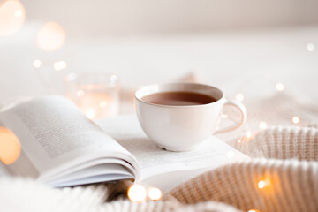 Open book with cup of coffee in bed with knit cloth sweater over glow christmas lights at...