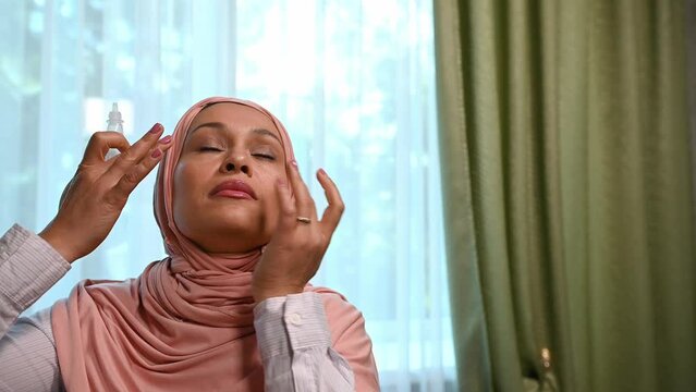 Close-up portrait Arab Muslim pretty woman with head covered in a pink hijab, using pharmaceutical eye drops, dripping into eyes to soothe irritation due to tension associated with overwork on laptop