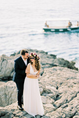 Fototapeta na wymiar Groom in sunglasses hugs bride standing on the rocks by the sea
