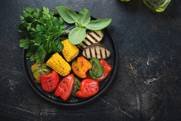 Round cast-iron tray with grilled vegetables and fresh parsley, above view on a dark-brown stone background, horizontal shot with space