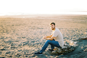 Fototapeta na wymiar Young man in jeans sitting on a snag on a sandy beach
