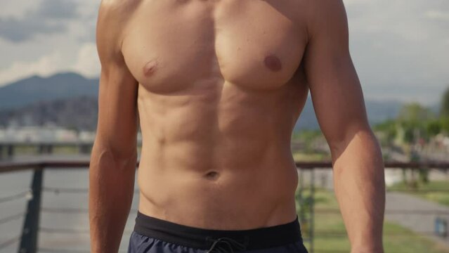 Athletic Caucasian man walking topless on the seashore after workout. Front view of young sportsman walking by wet sand on beach with elastic band. Man's beauty concept. High quality 4k footage. 