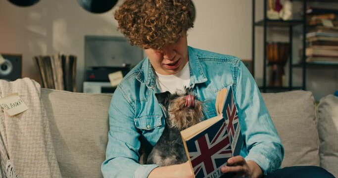 A boy with a dog on lap is reading an English dictionary, studying for a test, hugging the four-legged dog who is licking face. A teenager's love of animals spending time in the living room.
