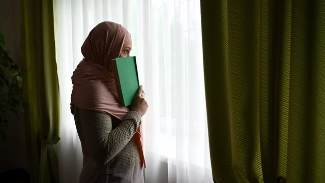 Beautiful confident Middle-Eastern Muslim pregnant woman in pink hijab, holding a holy book, whispering prayers while praying to god standing by a window at home interior
