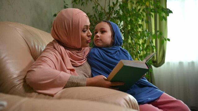 Beautiful Muslim woman in pink hijab, reading fairy tales to her adorable little daughter, sitting together on a comfortable armchair in the cozy home interior
