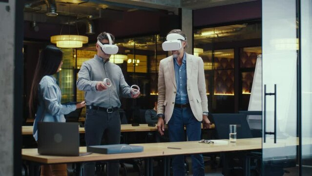 Multi-ethnic office workers testing VR headsets together, using tablet computer and wireless controllers, watching data, working in modern office of hi-tech company. Cyberspace digital technology