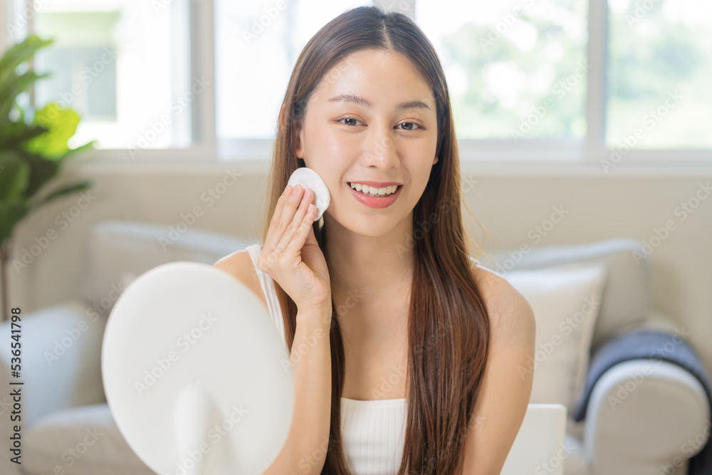 Wall mural Happy beauty, beautiful asian young woman, girl smiling, looking in mirror, holding cotton pad, applying lotion by wipe on her face, removing makeup before shower at home. Skin care routine people.