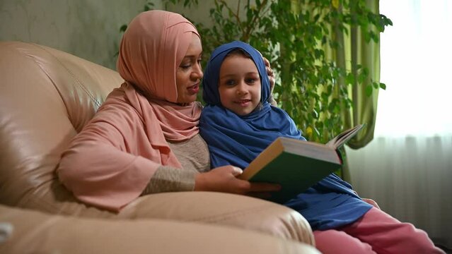 Portrait of a lovely caring Arab Muslim mother teaching her lovely daughter to read book. Happy family relationships. Woman and little girl are sitting together, hugging and discussing a fairytail.