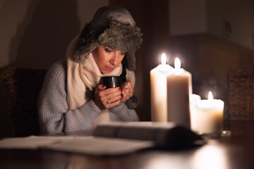 Freezing young woman in winter clothes warms her hands on cup of tea and lights with candles as...