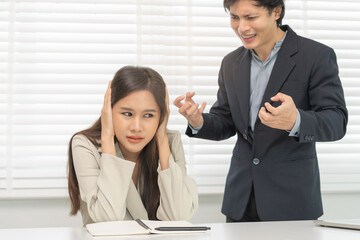 Angry boss, stressed asian young business, employee, secretary woman sitting at desk, hand cover her ears feel upset from annoying boss, colleague conflict with manager mistake about job at office.