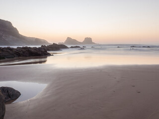 Fototapeta premium After sunset blue hour dreamy long exposure view of sand beach Praia da Zambujeira do Mar with rock and cliff and blurred ocean waves in pink light. Rota Vicentina coast, Odemira, Portugal