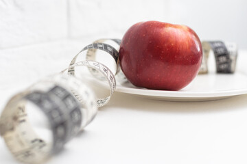 Slimming. A red-orange apple wrapped in a measuring tape lies on a white plate, on a white background. Calls to lose weight