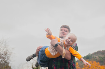 close-up portrait: dad plays with his little daughter, rolls her in his arms with an airplane in an autumn meadow on a warm day