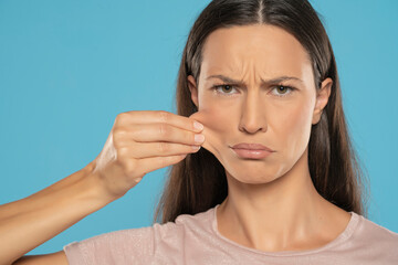Fototapeta premium Portrait of young unhappy brunette woman, pulling her cheek skin on blue background