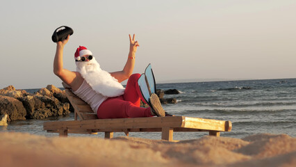Santa Claus sunbathes. Funny Santa Claus, in sunglasses and flippers, is sitting on lounger, on...