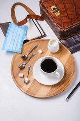 Black coffee in a white cup on a wooden tray on a white table next to a medical mask, notepad, pen, bag and magazine
