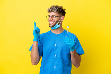 Dentist caucasian man holding tools isolated on yellow background pointing up a great idea