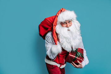 santa claus carrying huge bag of gifts on blue background while holding gift box in his hand.
