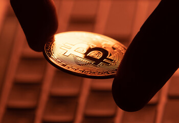 Golden coin with bitcoin cryptocurrency symbol in fingers with keyboard in the background in yellow light