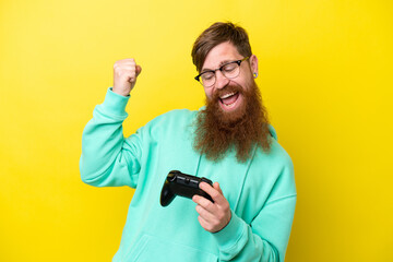Redhead man with beard playing with a video game controller isolated on yellow background celebrating a victory