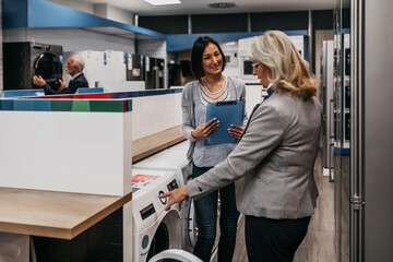 Saleswoman working in appliances store and showing to senior woman washing machines.