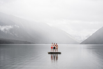 three caucasian boys together embracing standing dressed only in swimming trunks on the small platform of the calm and transparent lake between the mist that covers the sky and the green and leafy - Powered by Adobe
