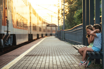 two little girls are sitting on a bench of the railway station and, out of fear of a loud sound passing by a train, they covered their ears with their hands so as not to hear - obrazy, fototapety, plakaty