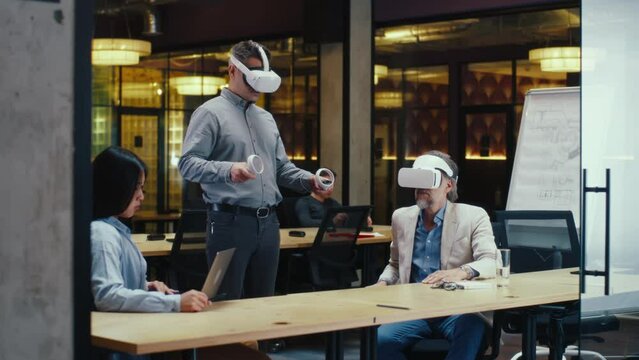 Multi-ethnic office workers testing VR headsets together, using tablet computer and wireless controllers, watching data, working in modern office of hi-tech company. Cyberspace digital technology