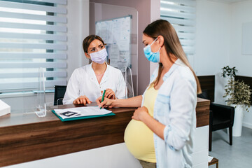 The pregnant woman is scheduling the next check-up with a gynecologist at the hospital. The nurse and pregnant woman are wearing protective face masks due to the coronavirus pandemic.