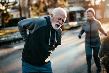 An older married couple jogging together outdoors. The wife helps her husband who injured his back while jogging.