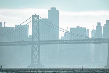 Idyllic view of Bay Bridge and urban skyline during foggy weather in San Francisco. Downtown district lying on sea front with cloudy sky in background. Famous tourist attraction in city.