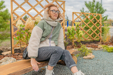 Young blonde woman in warm autumn clothes sits on wooden bench in recreation park and looks into distance.