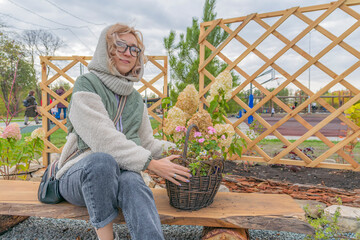 Young blonde woman in warm autumn clothes sits on wooden bench. Next to her is basket of flowers. Evening walk in fresh air.