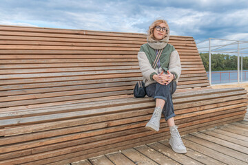 Young blonde woman in warm autumn clothes sits on wooden bench in leisure park and looks away.