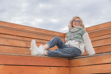 Young blonde woman in warm autumn clothes sits on wooden bench in recreation park and looks at camera.