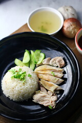 Hainanese chicken rice served on a black plate with dipping sauce and hot broth on white table.