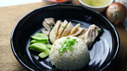 Hainanese chicken rice served on a black plate with dipping sauce and hot broth on white table.