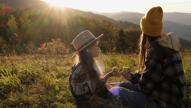 Beautiful young mother with pretty little daughter eating apple sitting in grass on background epic autumn mountains during sunset. Caring mom enjoys spending weekends with girl child in fall nature