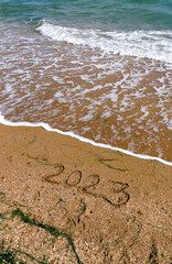 2023 written on the beach sand. Bright turquoise green sea waves with foam. Touristic concept for New year vacations. Template for touristic posters, postcards. Natural texture. Vertical orientation